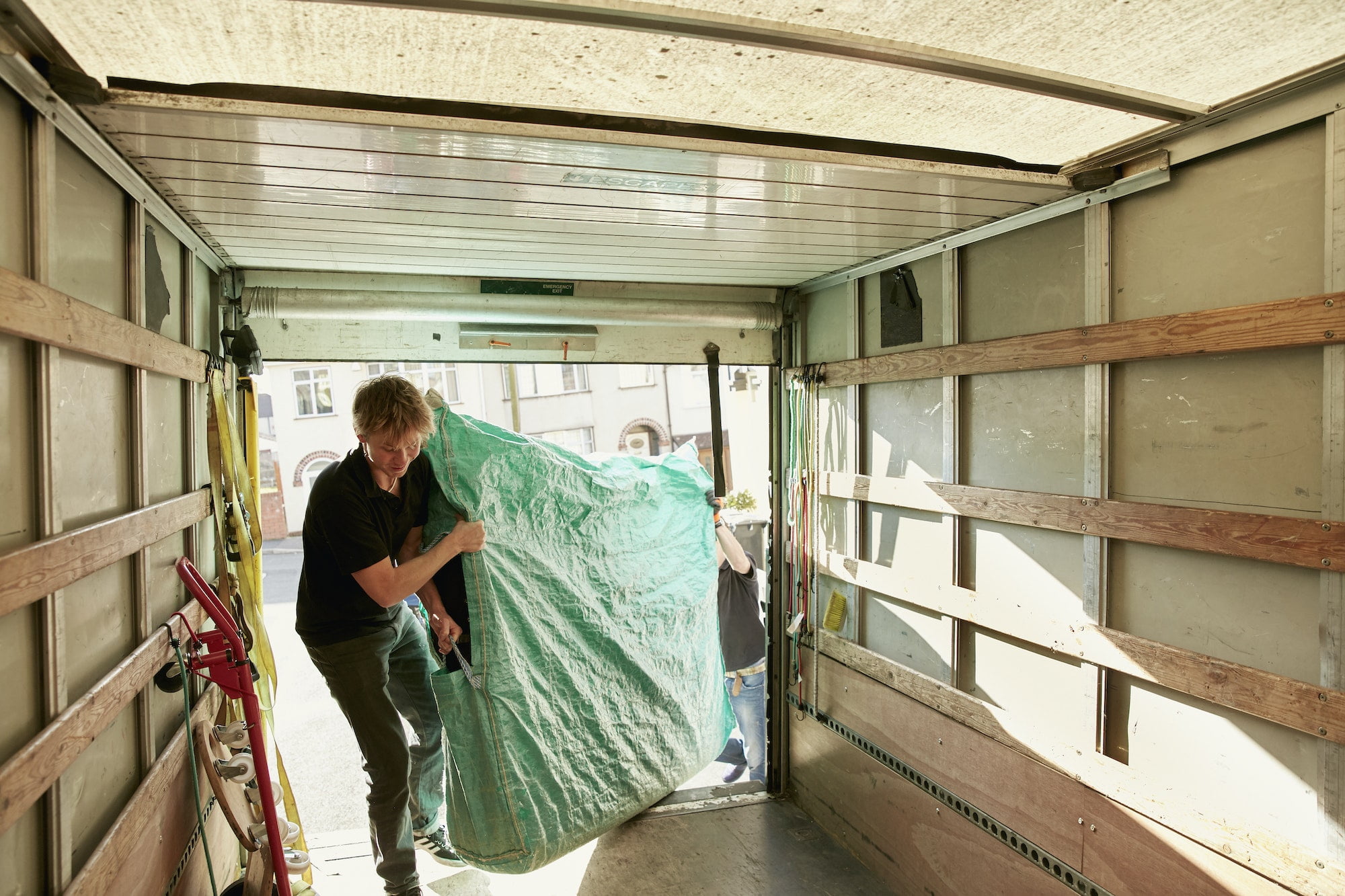 Removals business. A man lifting an item of furniture covered in green plastic into a removals van.
