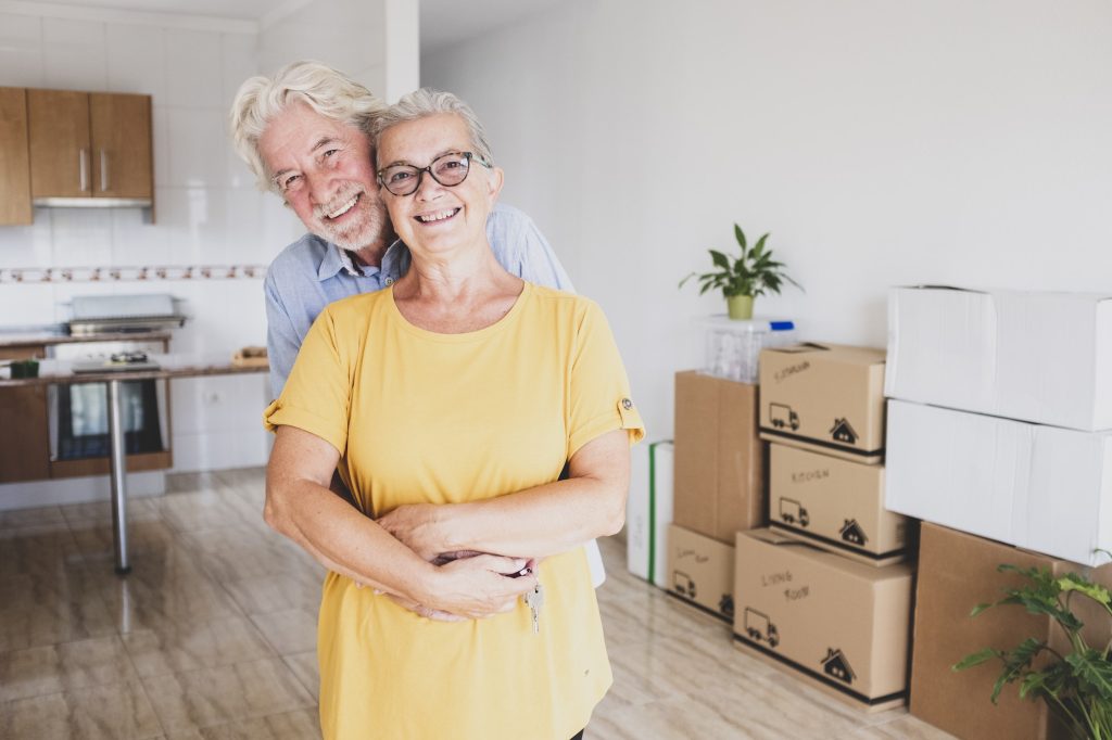 Smiling senior couple white-haired embraced during relocation, with moving boxes on the floor